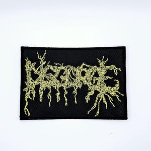 May include: Black rectangular patch with the word "AZATHOTH" embroidered in a yellow, stylized, and gothic font. The patch has a woven texture and is set against a white background.