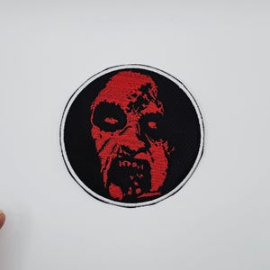 Puede incluir: Parche bordado con una cara de zombi roja sobre un fondo circular negro. La cara del zombi tiene detalles y está rodeada por un borde blanco. Ideal para ropa o accesorios.