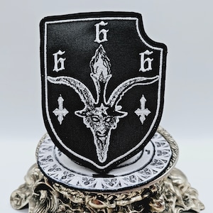 Op de afbeelding: Zwart-witte geborduurde patch met een geitenkop en drie 6's. De patch heeft de vorm van een schild en staat op een gouden standaard.