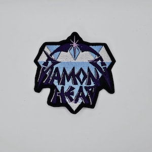 Könnte beinhalten: Gestickter Aufnäher mit den Worten "Diamond Head" in einer stilisierten Schriftart. Der Aufnäher hat eine Diamantenform mit schwarzem Rand, blauen und violetten Akzenten und einem hellblauen Hintergrund.