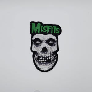 May include: Embroidered patch featuring the Misfits skull logo. The skull is white and gray with black accents, and the word "MISFITS" is embroidered in green above the skull. The patch has a black border.