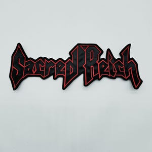 May include: A black embroidered patch featuring the text Sacred Reich in a stylized, angular font with a bold red border. The lettering is connected in a single piece, showcasing sharp, pointed edges and a distinct, heavy-metal aesthetic design.