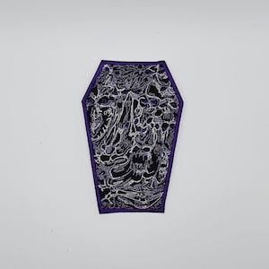 May include: A coffin-shaped patch with a black background and intricate white and purple embroidery. The patch has a purple border and features a detailed design of floral and skull-like elements, perfect for adding a unique touch to clothing or accessories.