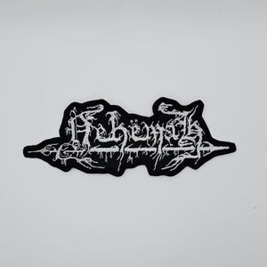 Puede incluir: Parche bordado en blanco y negro con la palabra "Behemoth" en una fuente gótica estilizada. El parche tiene un fondo negro con costuras blancas y una forma irregular. Está diseñado para su aplicación en ropa o accesorios.