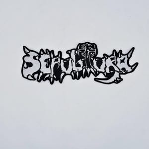May include: A black and white embroidered patch with the word "Sepultura" in a stylized, gothic font. The patch has a jagged, irregular border and is likely designed for application on clothing or accessories.