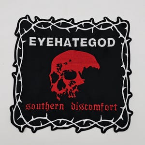 Puede incluir: Parche negro con un gráfico de calavera roja y el texto "EYEHATEGOD" y "southern discomfort" en blanco y rojo. El parche está enmarcado por un diseño de alambre de púas blanco.