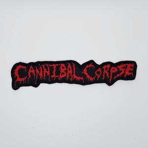May include: A black embroidered patch featuring the words "CANNIBAL CORPSE" in red, with a distinctive, stylized font. The patch has a rough, uneven border. It is designed to be sewn onto fabric. The background is a plain white.