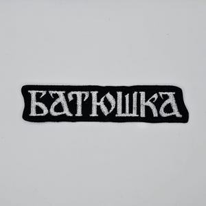 May include: A black embroidered patch with the white text "БАТЮШКА" in a stylized font. The patch has a rectangular shape with rounded corners. The text is centered on the patch.