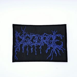 May include: Black rectangular patch with blue embroidered text. The text is stylized and appears to be a band logo or emblem. The patch has a black border and is set against a white background.