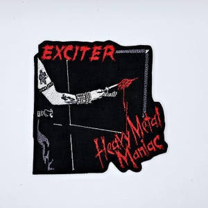 May include: Black embroidered patch with the word "EXCITER" in red, above a graphic of an arm holding a knife, with the words "Heavy Metal Maniac" in red. The patch has a black background.
