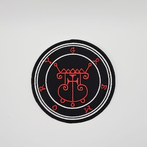Könnte beinhalten: Ein runder, schwarzer Aufnäher mit weißem Rand und rotem Stickdesign. Das Design zeigt ein zentrales Symbol mit aufwendigen Details und umgebenden Buchstaben. Geeignet für Kleidung oder Accessoires.