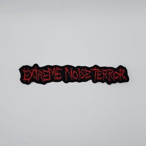 Puede incluir: Parche bordado negro con las palabras "EXTREME NOISE TERROR" en rojo, con letras dentadas. El parche es rectangular y tiene un borde negro. El texto está en negrita, con una fuente desgastada.