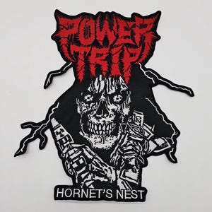 Pode incluir: Patch preto e branco com uma figura esquelética e o texto vermelho "POWER TRIP" no topo. As palavras "HORNET'S NEST" estão na parte inferior. O design tem uma estética punk rock.