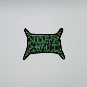 Może przedstawiać: Czarna naszywka z napisem "NUCLEAR ASSAULT" w kolorze zielonym, obramowana złotem. Naszywka ma unikalny kształt z ostrymi krawędziami. Tekst jest pogrubioną, stylizowaną czcionką, sugerującą przedmiot związany z zespołem lub muzyką.