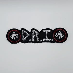 Może przedstawiać: Czarna naszywka z białym napisem "D.R.I." w stylu distressed. Naszywka ma logo biegnącej postaci w czerwonym okręgu na każdym końcu. Naszywka jest prostokątna o unikalnym kształcie.
