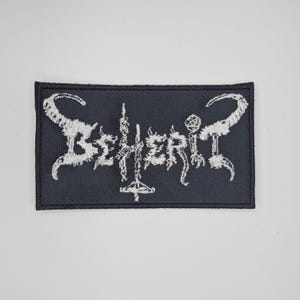 May include: A rectangular black patch with the white embroidered word "Beherit" in a stylized, gothic font. The patch has a black border and is likely for clothing or accessories.