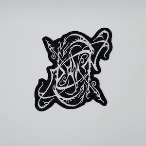 May include: Black and white embroidered patch with an abstract design. The patch features a black background with a white, swirling, and stylized text design. The patch has a unique, irregular shape.