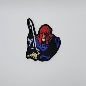 May include: Embroidered patch featuring a figure with a red mask and a sword. The figure is set against a black background with blue and gold accents. The patch is likely for clothing or accessories.