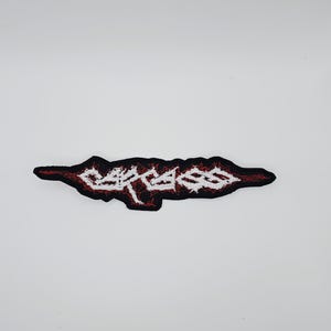 Puede incluir: Un parche bordado en negro y rojo con la palabra "CARCASS" en blanco, con una tipografía estilizada. El parche tiene una forma irregular y está diseñado para coserlo en ropa o accesorios.