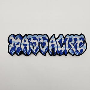 Puede incluir: Parche bordado con la palabra "SAVAGE" en letras de estilo graffiti azul y blanco. El parche tiene un borde negro y está sobre un fondo blanco. Las letras tienen un diseño único y estilizado.