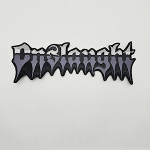 May include: An embroidered fabric patch featuring the word Onslaught in a stylised, jagged, black-letter font. The lettering has a gradient effect, transitioning from a light grey at the top to a darker purple-grey at the bottom, with a black border.