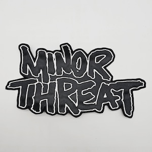 Peut inclure: Écusson noir avec l'inscription "MINOR THREAT" en lettres stylisées et audacieuses. Le texte est souligné d'une couture blanche, créant un fort contraste. Ce patch est probablement destiné aux vêtements ou accessoires.