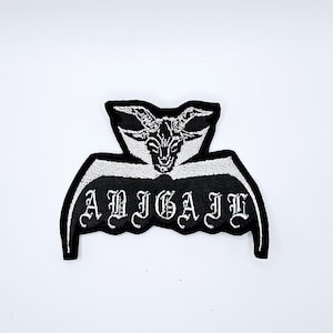 May include: Black and white embroidered patch featuring a goat head design with the name "ABIGAIL" in gothic lettering. The patch has a unique shape with curved edges and is likely for clothing or accessories.