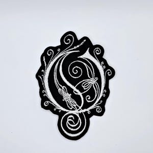 Peut inclure: Écusson brodé noir et blanc représentant la lettre "G" ornée de volutes et de motifs décoratifs. Le fond est noir, avec des coutures blanches contrastantes. Les bords sont festonnés.