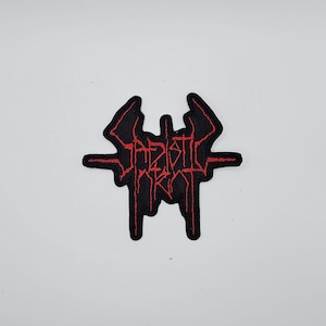 May include: A black patch with red embroidered text. The text is stylized and appears to be a band logo or emblem. The patch has a unique shape with a horizontal bar and vertical elements.