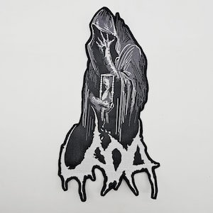 以下が含まれることがあります： 黒と白のパッチ。フード付きの人物が額縁に入った画像を持っています。その人物はマントをまとっており、下部には滴るような様式のテキストデザインがあります。このパッチはおそらく衣類やアクセサリー用です。