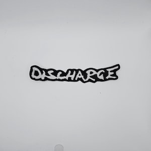 Puede incluir: Parche bordado en blanco y negro con la palabra "DISCHARGE" en una fuente estilizada. El parche tiene un fondo negro con letras blancas. El parche es rectangular y ligeramente curvado.