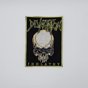 Op de afbeelding: Zwarte geborduurde patch met een schedelontwerp. Het woord "DEVASTATION" staat in gele, gestileerde tekst boven de schedel. Het woord "IDOLATRY" staat in gele tekst onder de schedel. De patch heeft een gele rand.