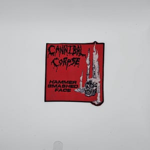 May include: A square, red patch with the words "CANNIBAL CORPSE" in black, gothic lettering. Below, the words "HAMMER SMASHED FACE" are printed in white. A black and white illustration of a skull and skeletal structure is on the right.