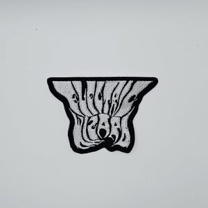 Puede incluir: Un parche bordado en blanco y negro con la palabra "SUGAR" en una fuente estilizada y ondulada. El parche tiene una forma única y abstracta con un borde negro, sobre un fondo blanco.