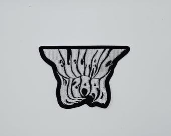 May include: A black and white embroidered patch with the word "SUGAR" in a stylized, wavy font. The patch has a unique, abstract shape with a black border and is set against a white background.