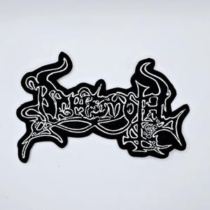 May include: Black and white embroidered patch featuring the word "Rebellion" in a gothic font. The patch has a black background with white stitching. The text is in a stylized, decorative font. Suitable for clothing or accessories.