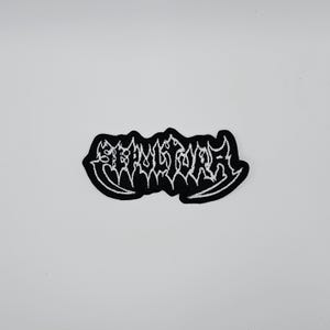 May include: A black and white embroidered patch featuring the word "SEPULTURA" in a gothic font. The patch has a black background, white stitching, and a white border.