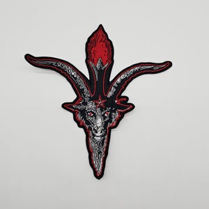 May include: A black and red embroidered patch featuring a goat's head with a star on its forehead. The patch has a red outline and a flame-like shape above the head. The goat's eyes are red.
