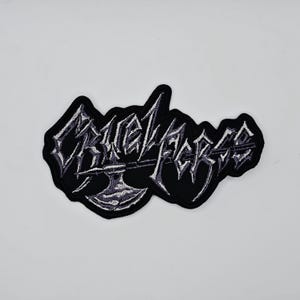 Puede incluir: Un parche bordado negro con el texto "CRUEL FORCE" en fuente gótica. El parche presenta un diseño de hacha gris debajo del texto. El parche tiene un borde negro, para ropa o accesorios.