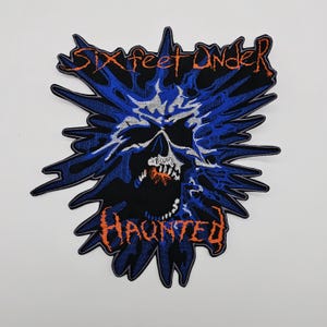 May include: Embroidered patch featuring a skull with an open mouth, surrounded by a blue and black burst design. The words "SIX FEET UNDER" and "HAUNTED" are in orange lettering.
