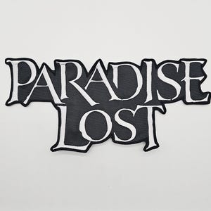 Puede incluir: Un parche bordado en blanco y negro con las palabras "PARADISE LOST" en una fuente estilizada. El texto blanco está delineado en negro sobre un fondo negro. El parche tiene una forma irregular única.
