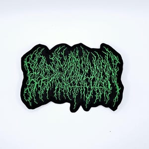 May include: Black embroidered patch with green text in a stylized, organic font. The patch has a black border and a textured appearance. The design is a bold, eye-catching statement.