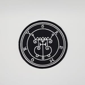 Könnte beinhalten: Ein runder, schwarzer Aufnäher mit einem weißen, gestickten Design. Das Design zeigt ein zentrales Symbol, das von einem kreisförmigen Rand mit stilisierten Buchstaben umgeben ist. Der Aufnäher ist für Kleidung oder Accessoires geeignet.