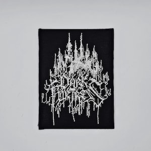 以下が含まれることがあります： 黒い長方形のパッチ。白い刺繍のテキストとゴシック様式の城のデザイン。テキストは、様式化されたフォントで「DARK POETRY」と書かれています。パッチは織り込まれたテクスチャで、衣類やアクセサリーに適しています。