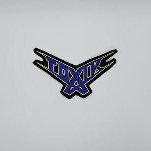 Pode incluir: Patch bordado com um design azul e dourado. O patch apresenta a palavra "TOXIC" em uma fonte estilizada, delineada em preto. O patch tem um formato angular único.