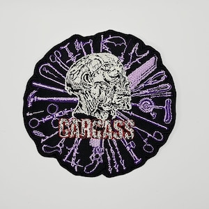 May include: A circular black embroidered patch featuring a central anatomical human head illustration in white and grey. Surrounding the head are purple embroidered surgical instruments. The word CARCASS is stitched in red and white lettering.