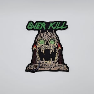 May include: Embroidered patch featuring the text "OVER KILL" in green above a detailed skull design. The skull has green eyes and is set against a black background with additional text at the bottom. The patch is likely for clothing or accessories.