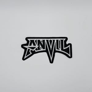 Op de afbeelding: Een zwart-wit geborduurde patch met het woord "ANVIL" in een gestileerd lettertype. De patch heeft een zwarte rand en een witte achtergrond voor de tekst, met een stervormig ontwerp aan de linkerkant.
