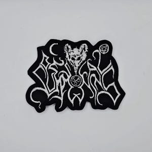 May include: Black embroidered patch with a white design featuring a horned skull, a crescent moon, and stylized lettering. The patch has a unique shape and is likely for clothing or accessories.
