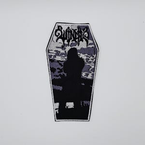 Op de afbeelding: Een doodskistvormige patch met het woord "SunnO)))" in gestileerde letters bovenaan. De patch heeft een zwart silhouet van een figuur in een doodskist, tegen een grijze en witte wolkachtige achtergrond.